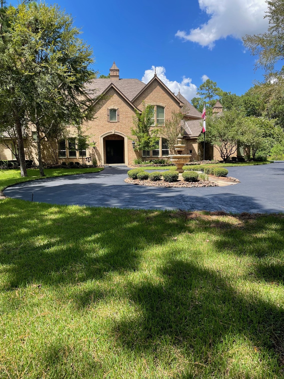 A tan brick house sits behind a paved circular driveway, an elegant fountain, and a green yard.